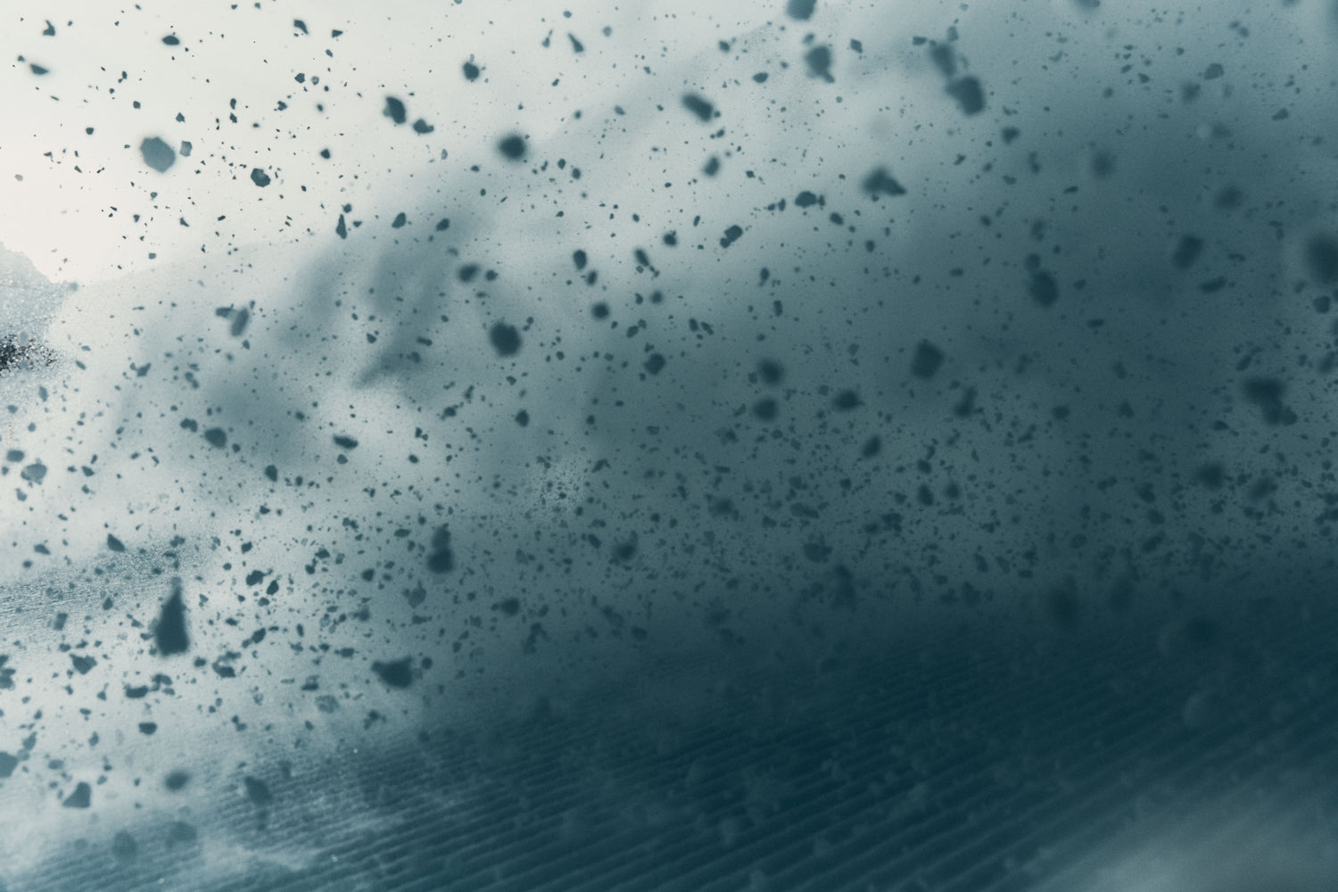 A close-up of a dark blue-gray textured surface covered with scattered black specks and smudges, with a blurred streak of light across the lower right area.